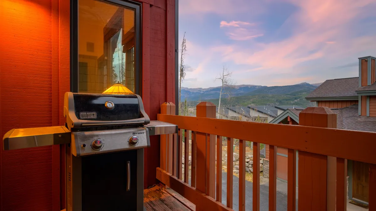 An exterior deck scene with a grill in the foreground, a sturdy deck railing, and sweeping mountain views. The setting is ideal for outdoor dining, barbecuing, and social gatherings, especially during sunset.
