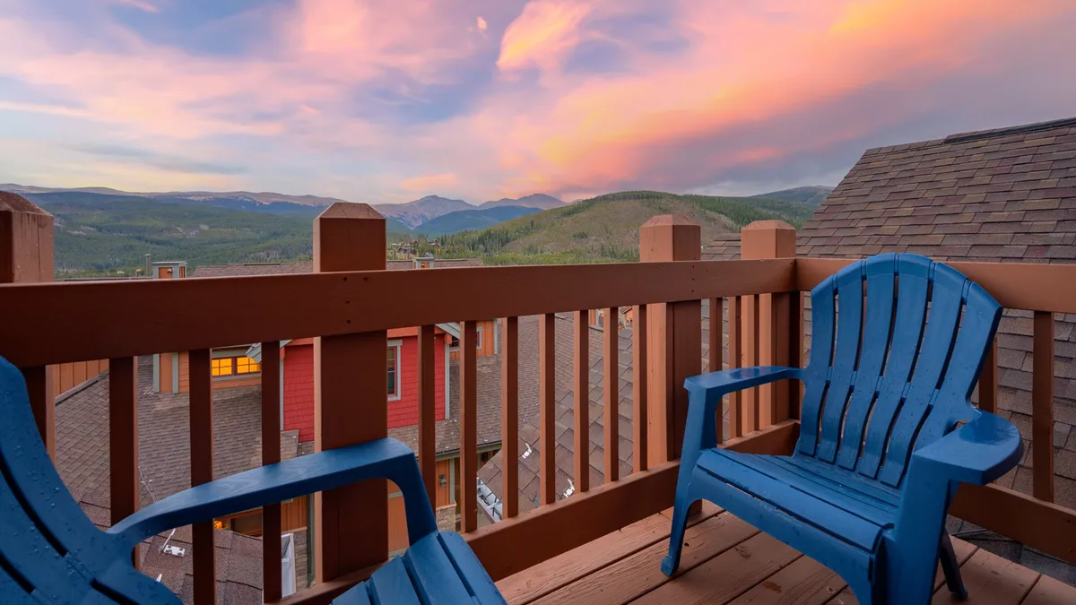 This image showcases a tranquil patio setting with blue Adirondack chairs overlooking a colorful Rocky Mountain sunset. The expansive mountain landscape provides a breathtaking backdrop, with the mood conveying relaxation, togetherness, and a profound connection to nature.