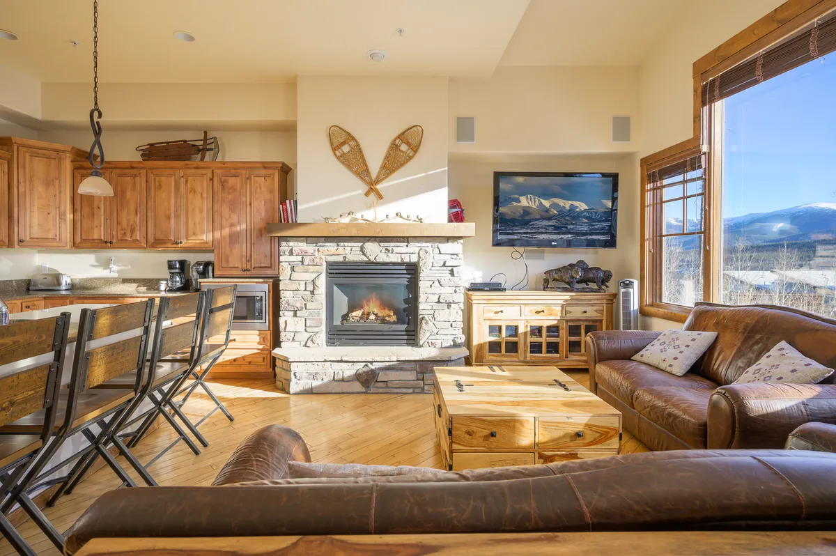 A spacious living area with a fireplace, kitchen island, and large windows overlooking the mountains. The arrangement of cozy seating, wooden elements, and open layout illustrates how the living and dining spaces flow together for gatherings and shared experiences.