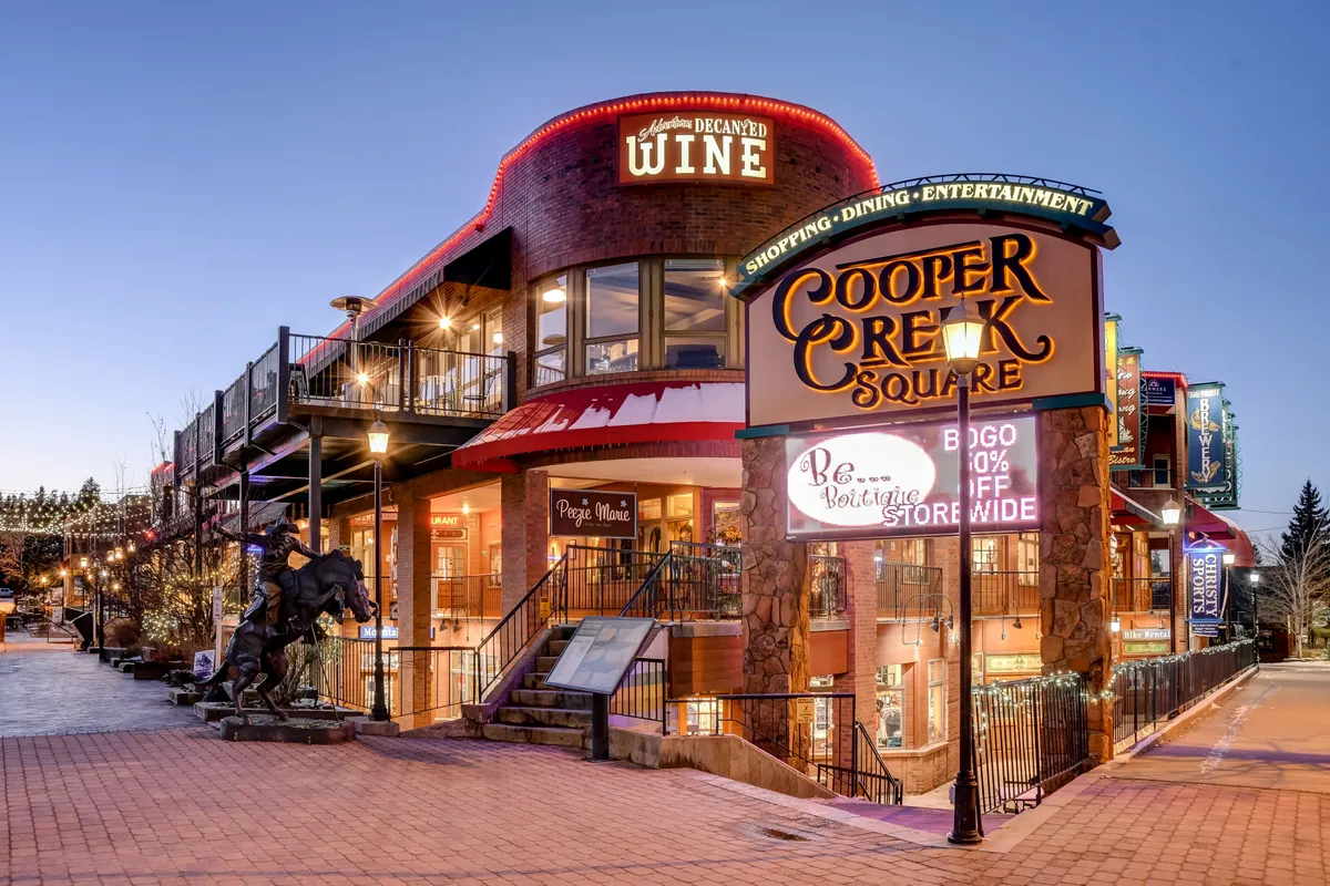 A vibrant exterior image of Cooper Creek Square, a centerpiece of downtown Winter Park, featuring illuminated signage, outdoor seating, and decorative lighting. The lively atmosphere invites groups to explore local dining, shopping, and entertainment just moments from the chalet.