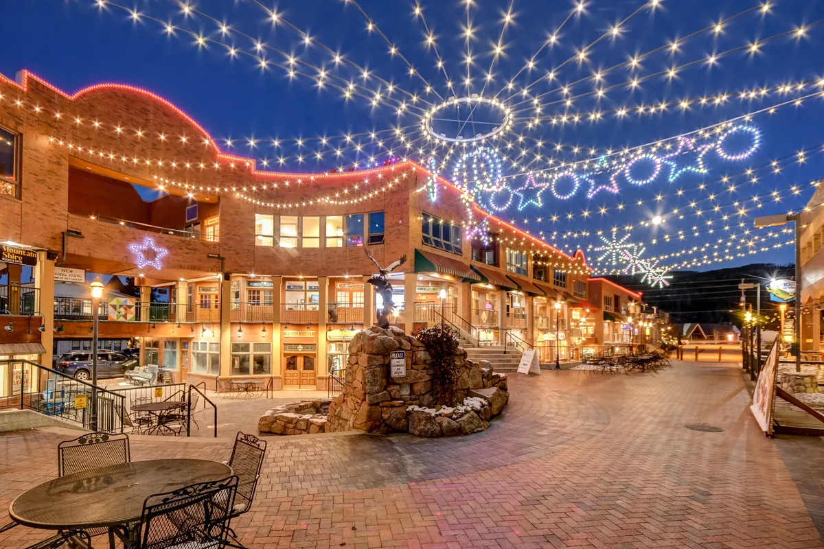An inviting exterior space adorned with twinkling string lights, decorative stars, a charming stone fountain, and a welcoming seating area. The mood is festive and communal, with people socializing, dining, and enjoying the lively atmosphere.