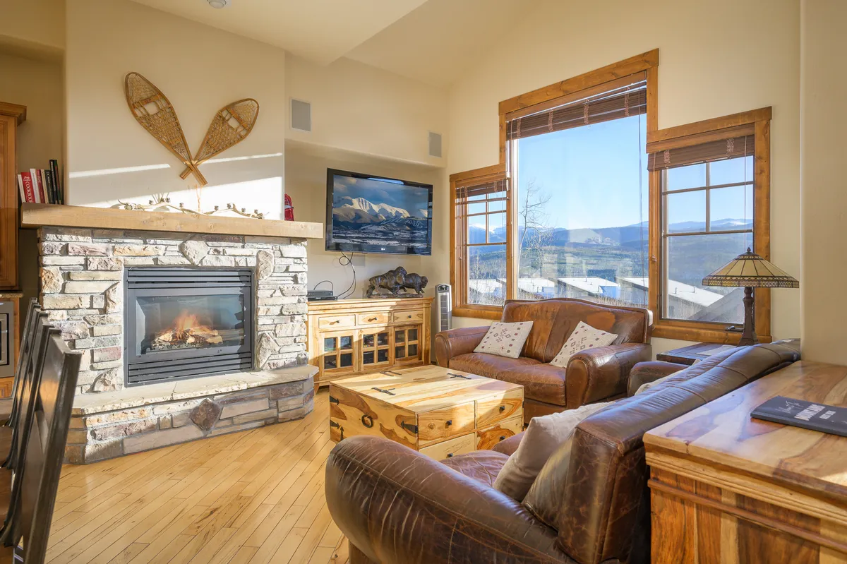 A warm and welcoming great room featuring a central fireplace flanked by comfortable seating, with large windows providing sweeping mountain views. Wooden furniture and soft textiles enhance the inviting atmosphere, and the space is perfectly designed for relaxing, watching TV, or gathering for games.