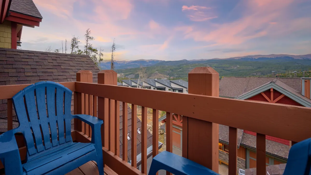 This image shows a serene exterior patio with blue Adirondack chairs arranged on a wooden deck, overlooking a stunning mountain view under a colorful sky. The scene suggests a cozy spot for groups to unwind, enjoy sunsets, and socialize with the dramatic natural landscape as a backdrop.