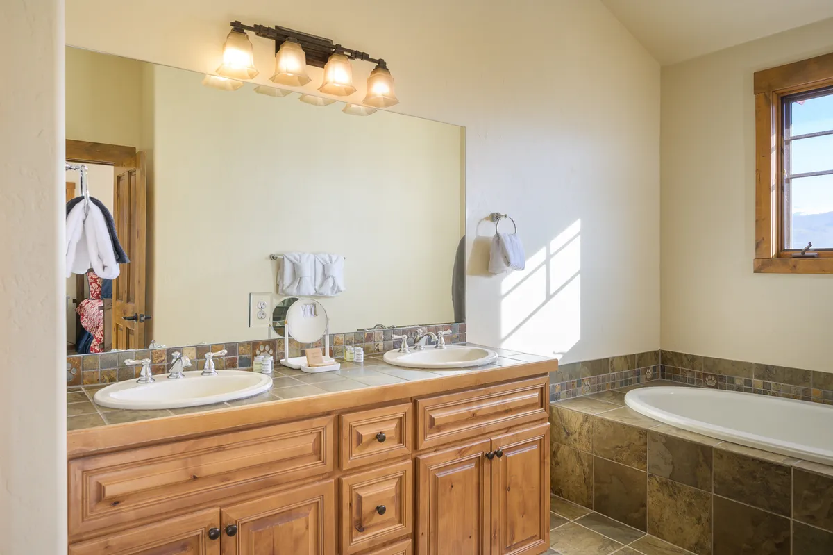 A spacious, light-filled bathroom with a double sink vanity, mirrored wall, and a bathtub set beside a sunny window. The elegant design and soothing atmosphere evoke the feel of a spa retreat.