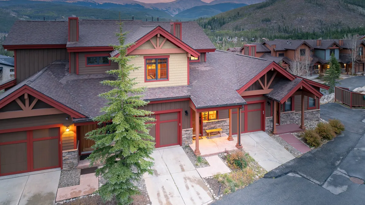 The image showcases the welcoming exterior of a luxury chalet, with a spacious front porch perfect for relaxing, elegant stone accents, floor-to-ceiling windows that invite in mountain light, and a beautifully landscaped garden. The setting suggests breathtaking sunset or starlit views, and a cozy, inviting atmosphere for gatherings.